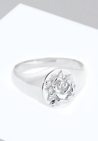Bague chevalière en argent avec une rose détaillée en relief et des feuilles sur la face ronde, présentée sur une surface blanche.