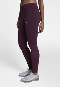 Kobieta w ciemnofioletowych legginsach Nike i jasnoszarych butach biegowych Nike, stojąca na tle jednolitego białego tła.