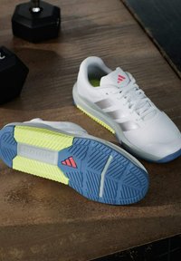 Białe buty sportowe z niebiesko-żółtymi podeszwami, jedno but stoi pionowo, drugie pokazuje wzór bieżnika, umieszczone na drewnianej podłodze obok hantli.