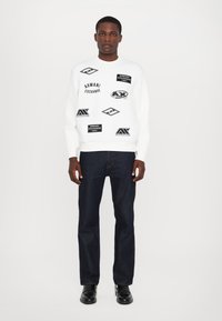 Biała bluza z czarnymi logo, okrągły dekolt i ściągacze, w połączeniu z ciemnymi dżinsami i czarnymi butami. Jednolity tło.