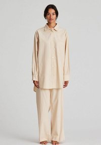 Camicia button-up in cotone beige con maniche lunghe e tasca sul petto, abbinata a pantaloni larghi dello stesso colore. Tessuto liscio.