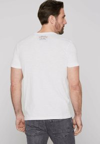 Weißes Baumwoll-T-Shirt mit kurzen Ärmeln, das auf dem Rücken am Halsausschnitt mit einem kleinen gedruckten Etikett versehen ist. Weicher Stoff, lockerer Schnitt, minimales Design.