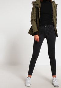 Nainen, joka pukeutunut oliivinvihreään parkatakkiin, mustaan poolopaitaan, tummiin kapeisiin farkkuihin ja valkoisiin lenkkareihin, seisoo jalat erillään yksiväristä taustaa vasten.