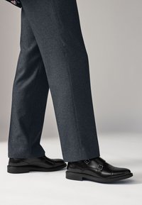 Chaussures en cuir noir à double boucle, texture contrastante avec un pantalon gris à motif à carreaux, doté d'un talon bas et d'un bout arrondi.