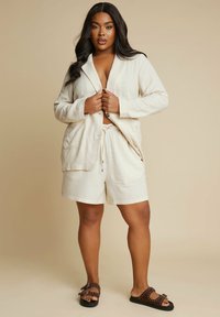 Kaksi osaa koostuva pellavapuku vaaleassa beessissä; mukana on kootut vyötäröshortsit ja väljät bleiserit, joissa on taskut; yhdistettynä ruskeisiin niittisandaaleihin.