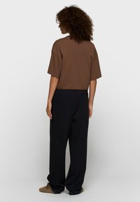 Persona con cabello rizado que lleva una blusa marrón holgada de manga corta, pantalones anchos negros y pantuflas marrones, de pie de espaldas sobre un fondo liso.