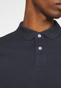 Polo bleu marine en tissu texturé, avec un col classique, une patte de boutonnage à deux boutons et un petit logo sur les boutons.