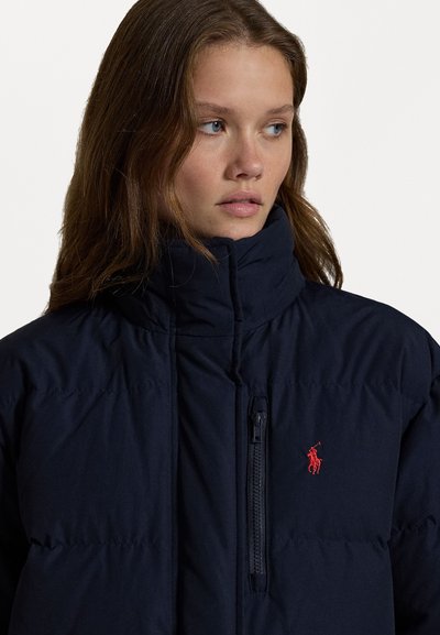 Jachetă puffer de culoare bleumarin cu guler înalt, având un fermoar frontal și un mic logo roșu pe piept. Textură netedă, design matlasat.