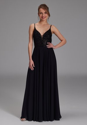 Frau in einem langen schwarzen Kleid mit einem glitzernden tiefen V-Ausschnitt und fließendem Rock, die mit einer Hand auf der Hüfte lächelt.