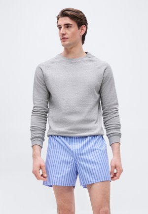 Mlajši moški, oblečen v svetlo sivo majico z dolgimi rokavi in modro-bele kratke hlače z navpičnimi črtami, gleda vstran.