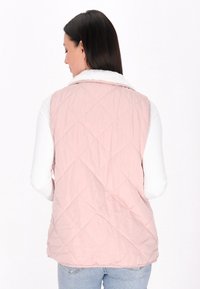 Rosa gesteppte Weste mit einem weißen Kunstpelzkragen, versehen mit Rautenmuster und ärmellosem Design, getragen über einem langen weißen Top.