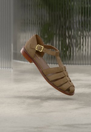 Brown leather sandal with multiple straps, gold buckle, and wooden sole, suspended above a light gray marble floor and textured glass background.