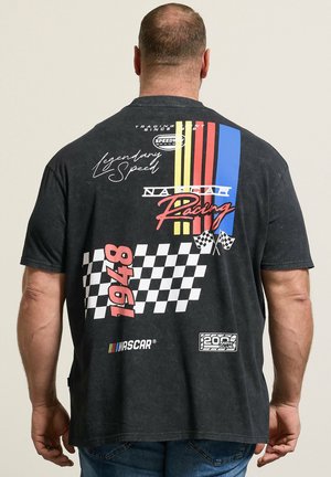 Mann trägt ein schwarzes NASCAR Racing T-Shirt mit Schachbrettflagge, vertikalen roten, gelben und blauen Streifen und dem Text "1948" auf dem Rücken.