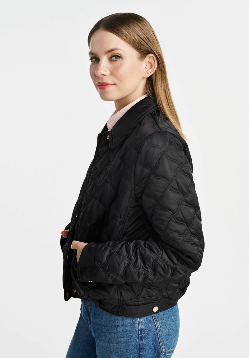 Veste matelassée noire avec un col, ornée d'accessoires en métal doré et de poches latérales. Portée sur une chemise rayée rose clair.