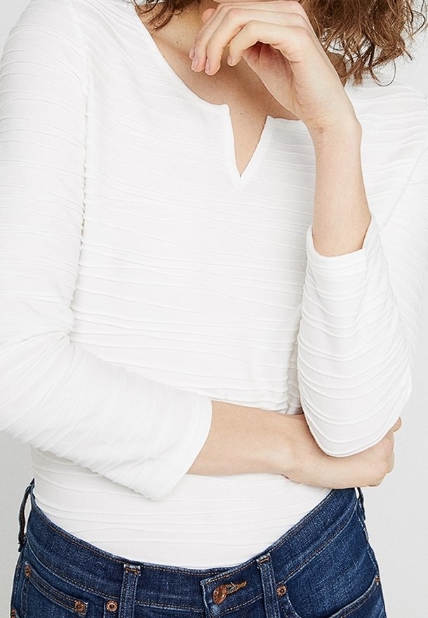 Top bianco con maniche lunghe e aderenti, caratterizzato da un leggero motivo texture e un piccolo scollo a V, abbinato a jeans di denim blu scuro.