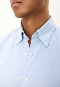 Lichtblauw overhemd met knopen, voorzien van een kleine kraag, witte knopen en een enkele knoopsgatdetail nabij de hals. Effen textuur.