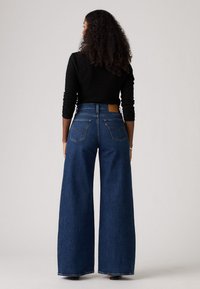 Jeans en denim bleu à jambe large avec taille haute, dotés d'une pièce en cuir à l'arrière et de deux poches arrière avec des détails de couture iconiques.