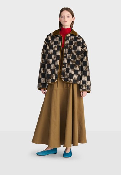 Femme portant une veste à carreaux noire et beige, un col roulé rouge, une longue jupe marron et des chaussures plates bleues, debout devant un fond uni.
