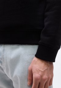 Κοντινό πλάνο του χεριού ενός ατόμου που ακουμπά δίπλα σε ανοιχτό μπλε τζιν, φοράει μαύρο πουκάμισο με μακριά μανίκια και ριμπ μανσέτες.