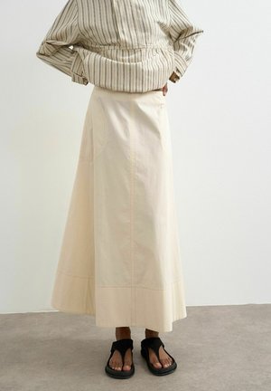 Person iført beige højtaljet A-formet nederdel, stribet langærmet skjorte og sorte åbne sandaler, står på gråt gulv mod en hvid væg.