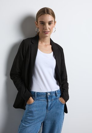 Blazer nero con un design sartoriale, abbinato a una canotta bianca a coste e jeans azzurri a vestibilità rilassata. Sono indossate semplici orecchini a cerchio.