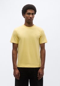 Κίτρινο βαμβακερό t-shirt με λαιμόκοψη και κοντά μανίκια. Διαθέτει μικρό λογότυπο στο στήθος. Φοριέται με μαύρο σορτς.