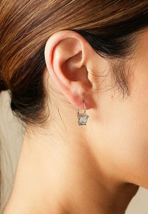 Silberne Creolenohrringe mit einem smaragdgeschliffenen, himmelblauen Stein, der im Ohrläppchen ruht, mit einer glatten, glänzenden Oberfläche.