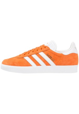 Baskets Adidas Gazelle en daim orange avec des bandes blanches, une semelle blanche et le texte "GAZELLE" sur le côté.
