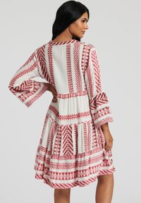 Vit klänning med röda geometriska mönster, klockärmar och ett lager på lager- bästa. Tillverkad av lättviktig tyg, som visar en strukturerad yta.