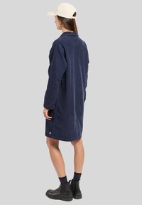 Robe chemise en velours côtelé bleu marine avec manches longues, poignets à boutons, fentes latérales et une étiquette logo discrète en bas. Portée avec une casquette claire et des bottes noires.