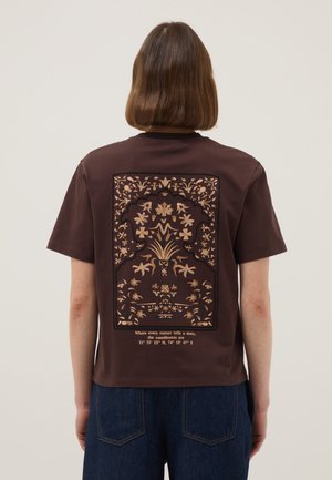 Persoon met kort bruin haar die een bruin t-shirt draagt met een beige bloemenpatroon en tekst inclusief coördinaten op de achterkant, gecombineerd met een blauwe spijkerbroek.