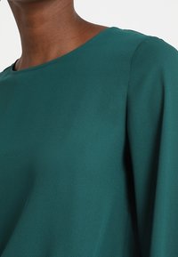 Blusa verde azulado con cuello redondo, fabricada en una tela suave y con mangas largas, que presenta una textura lisa y un ligero caimiento.