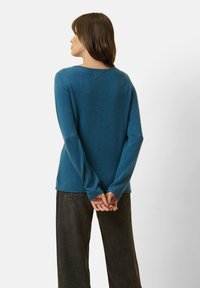 Suéter azul, suave, de manga larga con un escote redondeado, corte relajado y una textura especial. Combinado con pantalones oscuros de corte ancho.