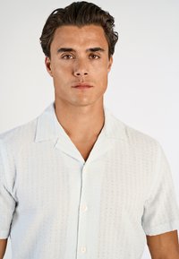 Jasnoniebieska koszula z krótkim rękawem o fakturowanym wzorze, z kołnierzem typu camp i zapinana na guziki z przodu. Wykonana z oddychającego materiału.