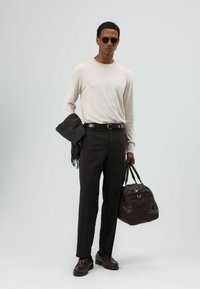 Beige shirt met lange mouwen, zwarte nette broek, donkerbruine leren schoenen, draagt een donkerbruine tas, met een grijze sjaal over één arm gedrapeerd.