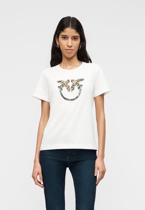 Nő fehér, rövid ujjú pólót visel, amely színes flitteres madármintával díszített, valamint sötétkék farmert, és világos, egyszínű háttér előtt áll.