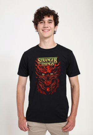 Maglietta nera di cotone con una grafica vivace in rosso e arancione di un volto con occhi luminosi, con la scritta "STRANGER THINGS" in verde sopra.