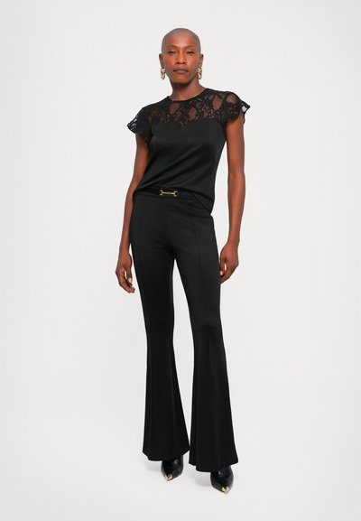 Personne portant un haut en dentelle noire, un pantalon évasé noir avec une ceinture à boucle dorée et des chaussures noires à bouts pointus, se tenant contre un fond blanc uni.