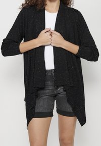 Svart cardigan med texturerad yta och flerfärgade prickar, med öppen framsida och långa ärmar, bärs över en vit topp och shorts.