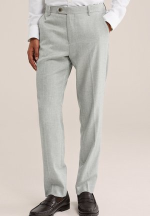 Homme portant un pantalon de costume gris clair, une chemise blanche et des mocassins en cuir brun foncé, debout avec une main dans la poche.