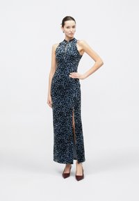 Donna in piedi con una mano sui fianchi, indossa un abito lungo senza maniche con stampa leopardata blu e nera, con spacco laterale e décolleté bordeaux a punta.