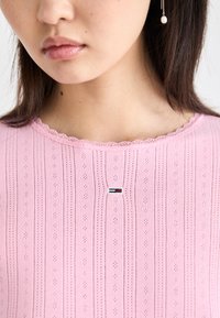 Lyserød top med struktureret ribbet mønster, bølget halsudskæring og lille logo detalje med røde og blå striber i midten.