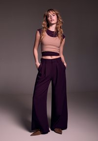 Top corto beige a coste abbinato a pantaloni viola a gamba larga e scarpe marroni con punta a punta. L'outfit presenta una silhouette aderente e rilassata.