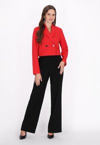 Blazer rosso corto con design doppiopetto, pantaloni neri a vita alta, dettagli a forma di cuore argentati e tacchi alti neri.