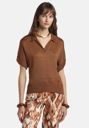 Ung kvinde iført en brun kortærmet striktop, dyreprintede bukser med vidde i benene, guldkæde og matchende armbånd, stående foran en hvid baggrund.