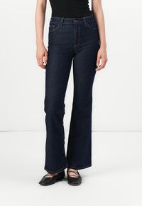 Γυναίκα που φοράει σκουρόχρωμο μπλε ψηλόμεσο τζιν μποτκάτ, μαύρη μπλούζα και μαύρες μπαλαρίνες με διασταυρούμενες λωρίδες, στηρίζεται σε λευκό φόντο.