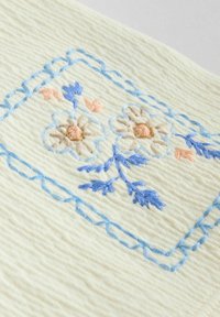 Tissu brodé avec un fond crème comportant des motifs floraux en bleu doux, rose clair et beige, bordé par un rectangle cousu en bleu.