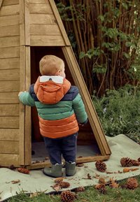 Padded detská bunda s pruhmi v zelenej, modrej a hrdzavej farbe; detail medvedej tváre na kapucni. Temné nohavice a zelené gumové čižmy. Pozadie z drevenej chaty.
