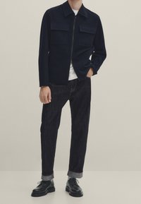 Tummansininen vetoketjullinen takki, jossa on kaksi rintataskua, käytetään valkoisen T-paidan päällä. Yhdistetty tummaan farkkuihin ja mustiin nauhakenkiin.