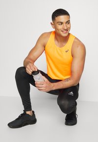 Camiseta deportiva de color naranja brillante con diseño perforado, combinada con leggings negros. La persona sostiene una botella de deporte transparente. Zapatillas deportivas negras.